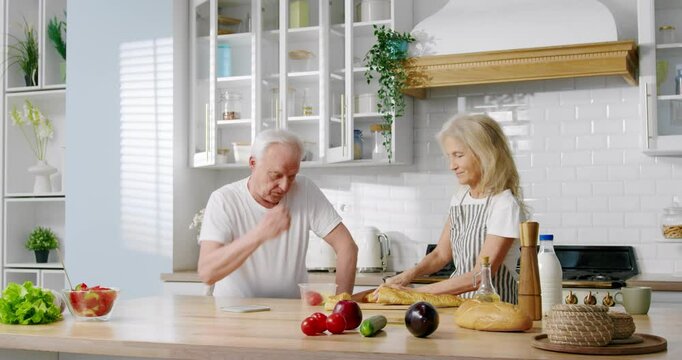 Aged spouses chatting at kitchen, woman cutting fresh bread, cooking dinner. Happy married couple, calm weekend at home with cozy interior, cheerful wife and grumpy husband, cute grandma and grandma