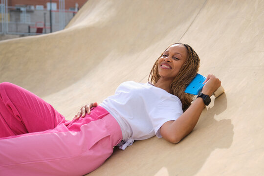 Stylish woman relaxing on skate ramp holding penny board and smiling