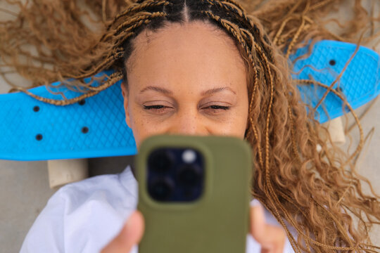 Young woman with blonde braids lying on skateboard using phone