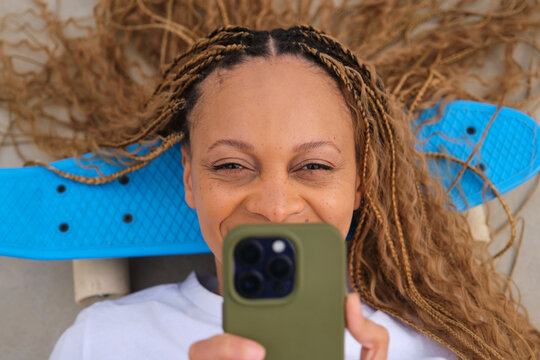 Young woman smiling and using smartphone lying on blue skateboard