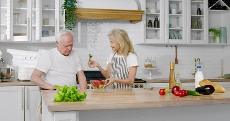 Happy family at kitchen, elderly woman cooking salad and husband refusing to eat . Healthy nutrition for lose weight and stay active in old age, vitamins in organic vegetables from eco farm for health