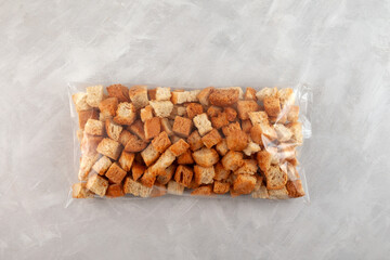 Crunchy bread croutons in transparent plastic bag on grey background. Pack of rustic homemade rusks. Wheat dehydrated bread, perfect topping for salads and mashed soups