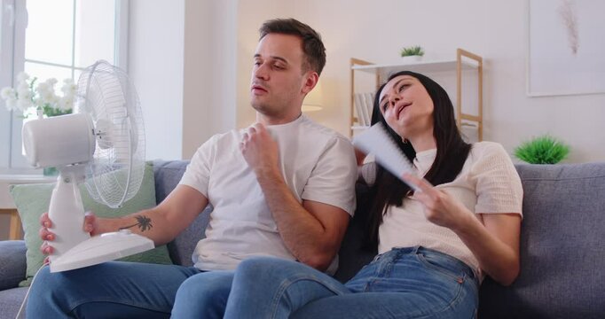 Couple with broken air conditioning system refreshing by fan on very hot summer day. Tired husband and wife sitting on couch at home enjoying fresh air blowing from electric fan. Summer heat concept