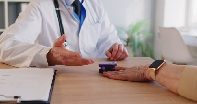 Unknown doctor measuring saturation with fingertip pulse oximeter to patient who came at medical examination in clinic, posing at table in cabinet. Measuring oxygen level in blood.