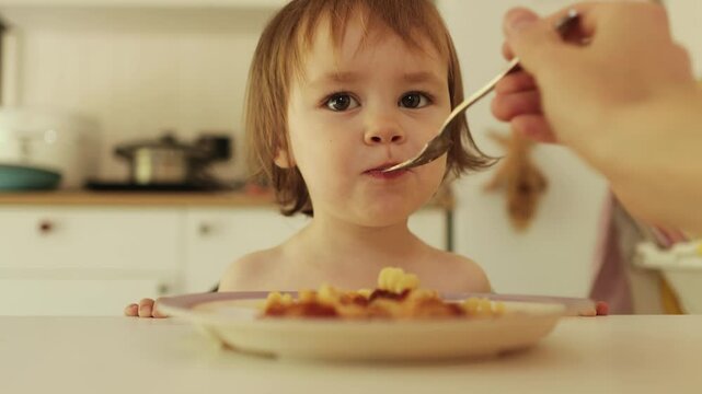 3 year old child happily eats pasta while sitting at a white table in a bright room, mother feeds the child with a fork. close-up of the hand