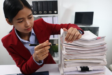 A young professional woman, examining a tall stack of documents with a focused expression.background are filled with more documents and black binders, suggesting a busy office environment