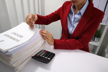 A young professional woman, examining a tall stack of documents with a focused expression.background are filled with more documents and black binders, suggesting a busy office environment