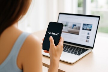 Woman using smartphone for online payment with laptop on desk in a bright home setting.