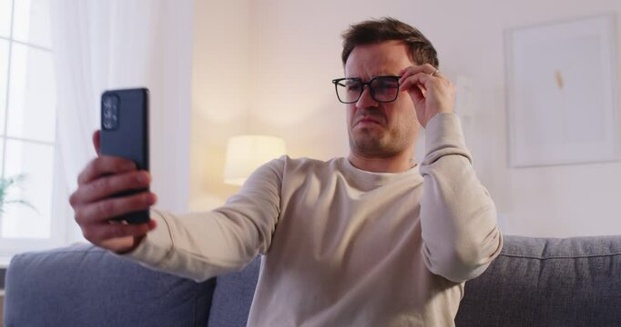 Young man wearing eyeglasses trying to read message on his smartphone having farsightedness eye disease. Male person with presbyopia looking at mobile phone screen. Vision problems concept. 4k video.