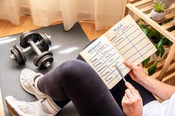 Woman sitting comfortably on a chair near a fitness mat and dumbbells, reviewing a workout plan while scheduling fitness activities for a balanced and healthy lifestyle