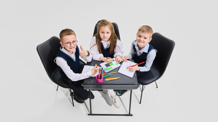 Two boys and girl in school uniform smiling and looking up from notebooks while sitting together at desk. Concept of academic joy, friendship, collaboration and shared educational journey.