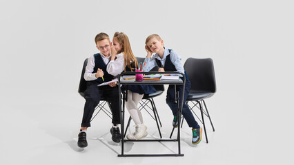 Girl whispering into boy's ear while seated at shared desk with pencils and notebooks as third classmate reacts. Concept of school gossip, distraction, fun and human interaction in learning.