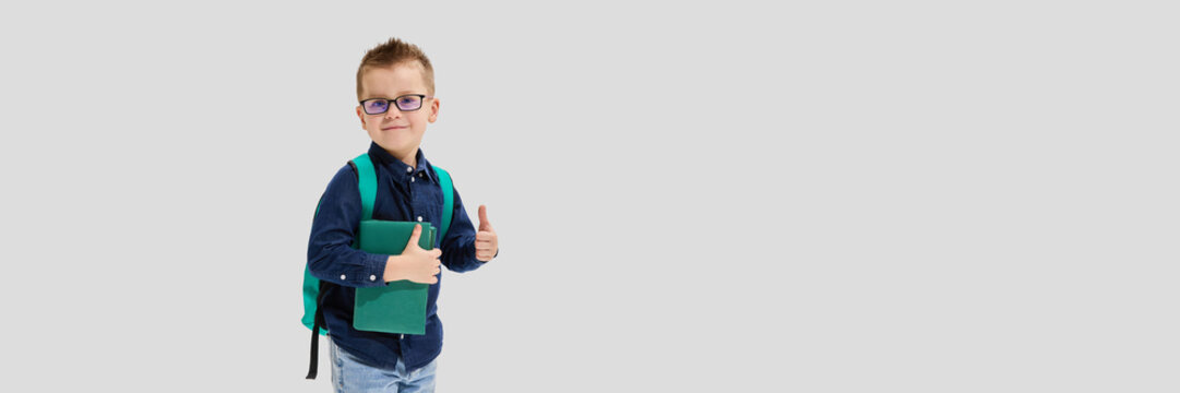 Banner. Smart boy with glasses in dark blue shirt holding green book and showing thumbs up against white background. Concept of confident student, good grades, school pride, knowledge, classroom. - Powered by Adobe