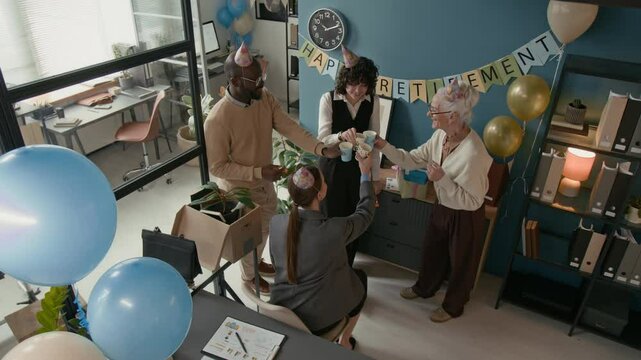 From above shot of cheerful diverse office team raising their cups in toast to celebrate senior woman retirement, hugging all together warmly, surrounded by balloons and festive decor