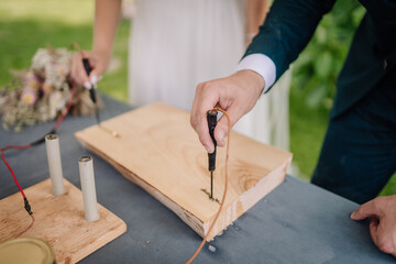 Close-up of two people woodburning on a wooden plank using pyrography tools, outdoors on a table. A bouquet and copy space are visible..
