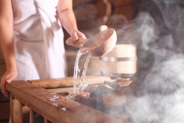 Woman pouring water onto stones surrounded by steam in sauna, closeup