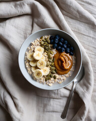 Fototapeta premium Creamy Oats for Better Sleep: Topped with Banana, Almond Butter, Pumpkin Seeds, Blueberries, Soft Beige Linen, Relaxing Evening Meal, Soft Light