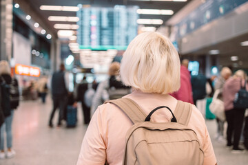elderly woman stands in the airport, looking up at the departure board, surrounded by fellow travelers. She appears to be checking her flight details, preparing for her upcoming journey.