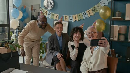 Cheerful senior retiree snaping selfie with her younger coworkers during retirement party, capturing joyful moment surrounded by smiles, balloons, and party hats