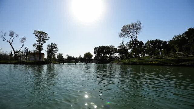 A drone on the water of a city lake in a park surrounded by trees. View of the promenade bridge. Cloudless summer day