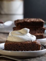 A slice of chocolate brownie with whipped cream. A stack of brownies in the background. A rustic-style table.