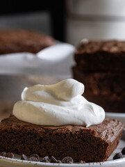 A slice of chocolate brownie with whipped cream. A stack of brownies in the background. A rustic-style table.