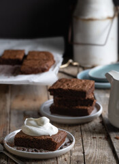 A slice of chocolate brownie with whipped cream. A stack of brownies in the background. A...