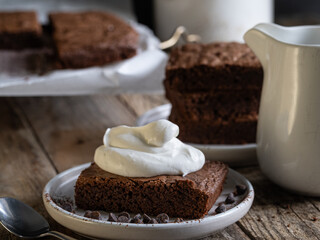 A slice of chocolate brownie with whipped cream. A stack of brownies in the background. A rustic-style table.