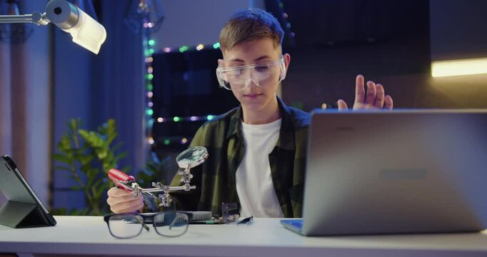 Boy in protective glasses soldering panel using soldering iron during revision training video on laptop in the evening at home