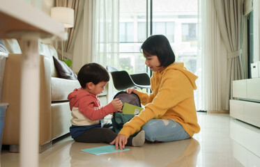 Fototapeta premium Asian young child and parent sit on the floor, packing a backpack with school supplies in a living room at home.