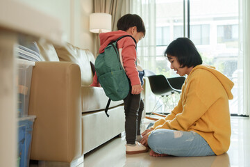 Fototapeta premium Asian young child with backpack stands near a sofa as their mother assists with tying shoes in a living room at home.