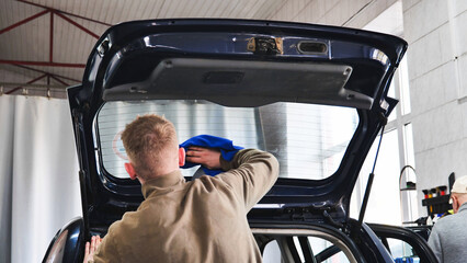 Car wash employee cleaning rear window of vehicle with open trunk