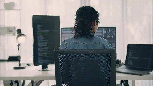 Rear view of female coder working on programming and UI development across multiple monitors in a modern tech office. Concept of multitasking and software engineering.