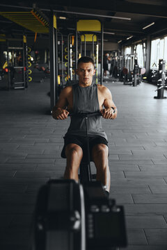 Vertical foto of strong young fit male sitting and using cable pulling machine, working out in modern gym. Handsome athlete man training on rowing machine at fitness center.
