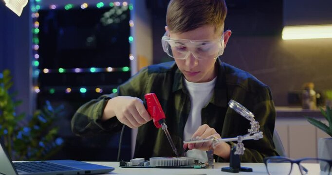 Boy in protective glasses sitting at the desk soldering on electronic panel at home in the evening.