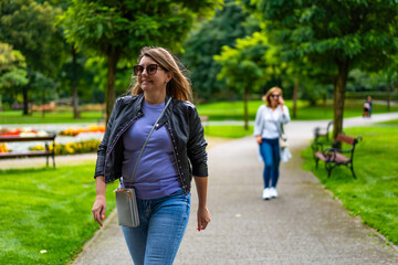 Obraz premium Two beautiful mature women walking separately, one goes in front, other in back, in city park. Front view