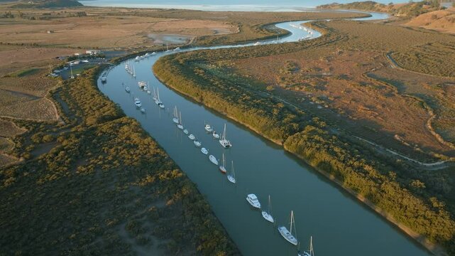 Aerial view of sailboats moored along a river in a rural landscape. The boats are lined up, reflecting in the calm water. Taken in New Zealand. CLEVEDON, NZ
