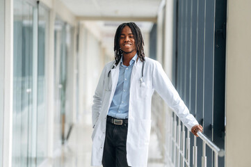 Positive facial expression, standing. Young male black doctor is in the corridor