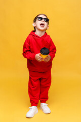 Cool little boy 3-4 years old in red track sport suit and black sunglasses holding yellow paper cup in hand on yellow background. Take away coffee concept. Copy space. High quality photo