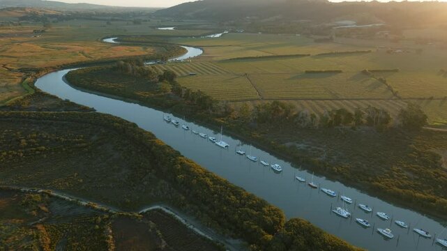 Aerial view of boats moored in a river, surrounded by lush greenery and farmland, bathed in the warm glow of the setting sun in New Zealand. CLEVEDON, NZ
