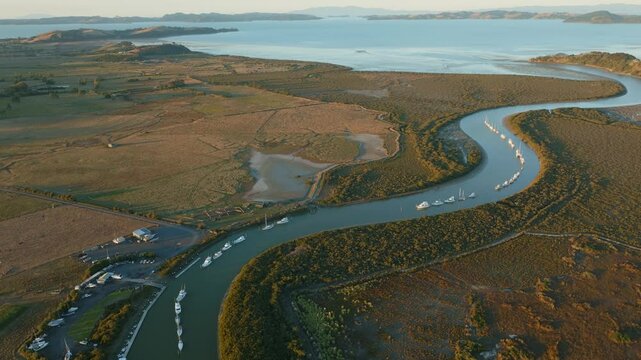 Aerial view of boats moored in a river, surrounded by lush greenery and mudflats, leading to the ocean in New Zealand. The scene captures the serene beauty of coastal life. CLEVEDON, NZ