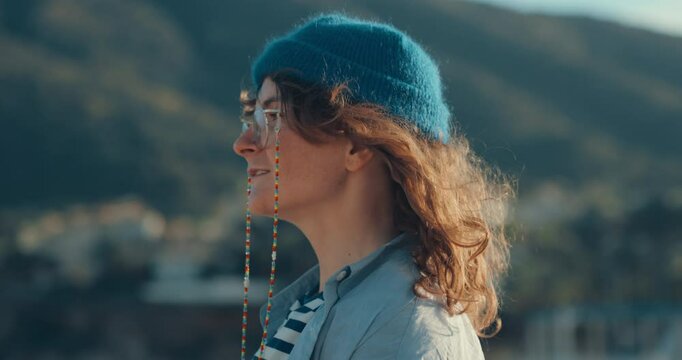 Authentic young woman in blue themed outfit, striped t-shirt, hand knitted blue wool beanie and prescription glasses and curly hair looks at ocean and then directly at camera. Candid image of youth