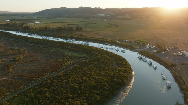 Aerial view of boats moored in a river, surrounded by lush greenery and farmland, bathed in the warm glow of the setting sun in New Zealand. CLEVEDON, NZ
