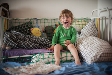 Naklejka premium A young child wearing a green shirt sits on a bed with various pillows and toys, laughing happily. The room is bright and inviting, creating a playful atmosphere.