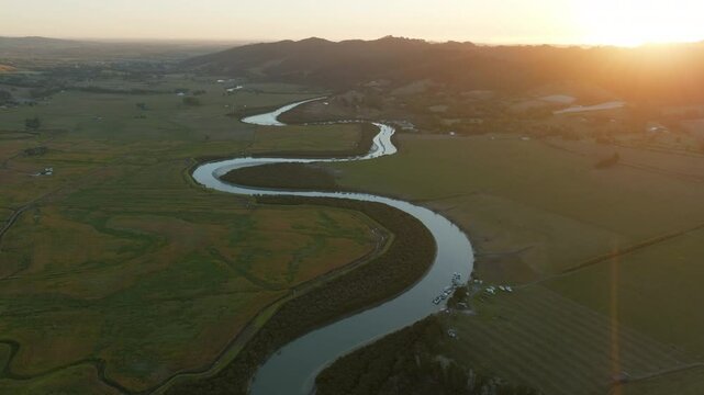 Aerial view of boats moored along the riverbank at sunrise in New Zealand. The boats are docked for recreational use and transport. The sun rises over the landscape. CLEVEDON, NZ