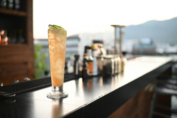 A tall glass of iced cocktail garnished with lime slices sits on a sleek rooftop bar counter, with a sunset city view in the background