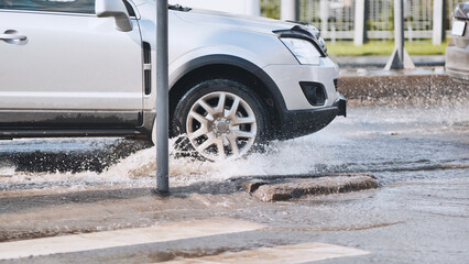 Silver sedan splashing water while driving through urban street puddle, wet asphalt reflecting...