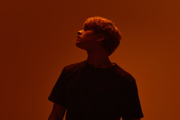 Silhouette of young man with sharp shadows and raised gaze in warm neon light against gradient...