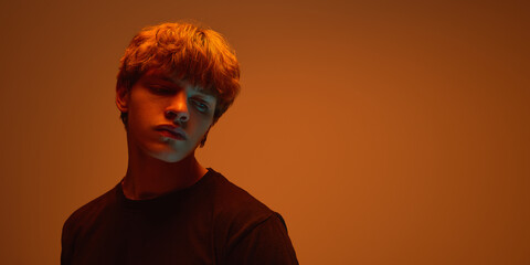 Poster. Young man with lowered gaze, dressed black t-shirt posing against warm orange background in bright neon light. Copy space. Concept of beauty, emotions, aesthetic, trends. Ad