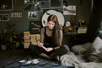 Beautiful goth teenager shuffling deck of tarot while sitting on bed with jet black wall decorated...